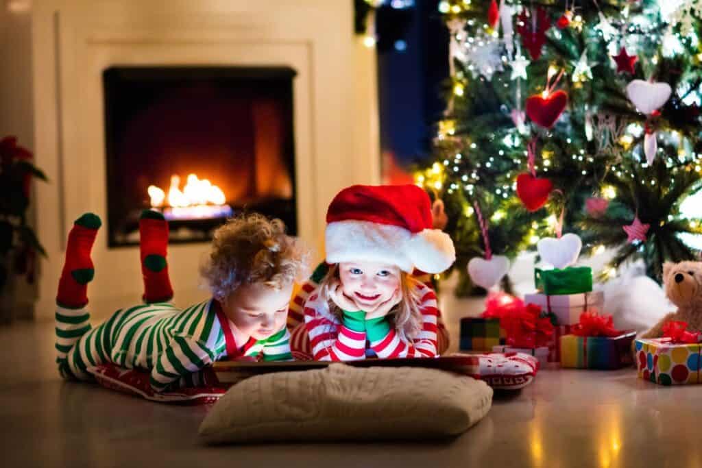 Two preschool girls looking at books near a Christmas tree with the light dim and fire going in the fireplace