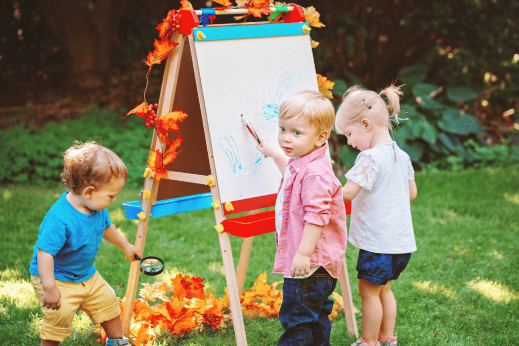3 preschool age children out side with and art easel. one boy has a magnifying glass, one boy is at the easel with a marker and the girl is standing next to him. 